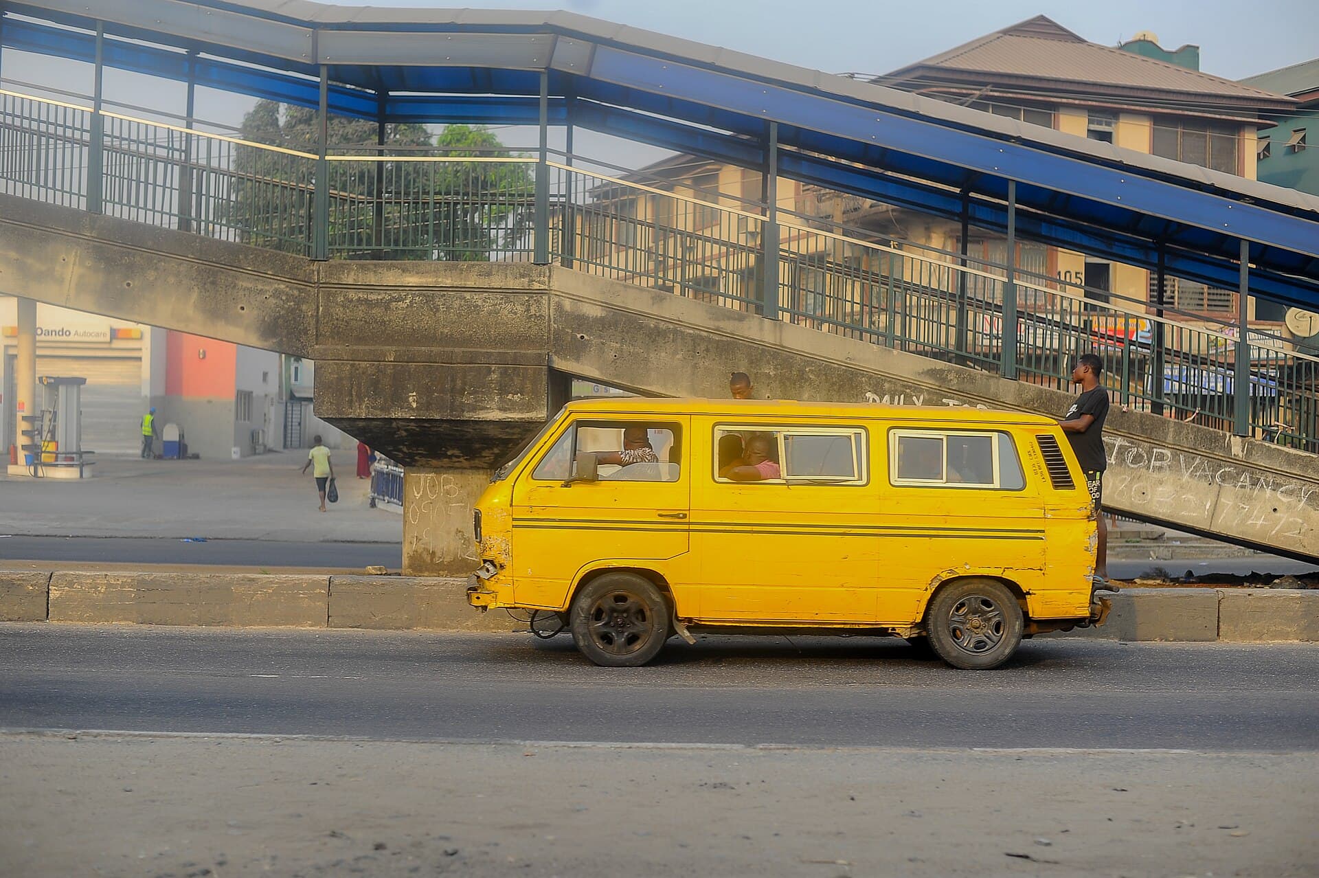 A yellow danfo bus pulled up beneath a Lagos pedestrian bridge at dusk